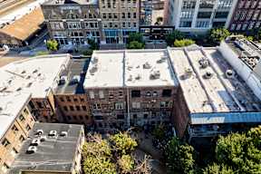 A large, multi-story building with a flat roof is surrounded by other buildings.