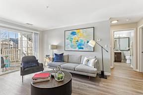A living room with a grey couch, a black chair, a coffee table with a red book and a basket on it, and a painting on the wall.