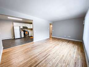 A room with wooden flooring and a kitchen in the background.