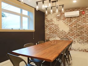 A long wooden table with chairs is in front of a brick wall with a window.