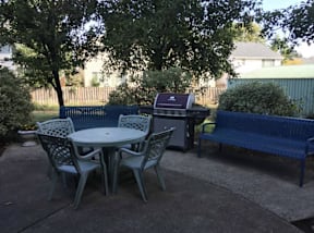A blue bench sits next to a table with chairs and a grill on a patio.