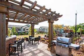 A wooden pergola over a patio with a grill and chairs.