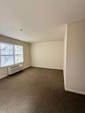 A room with a carpeted floor and a window with an air conditioning unit underneath it.
