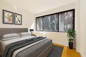 A bedroom with a bed, two pictures on the wall, a potted plant, and a window with a view of a building outside.
