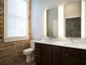 Bathroom with double vanity, backlit mirrors and exposed brick wall