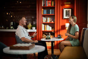 A man and a woman are having a conversation in a cozy room with a bookshelf in the background.