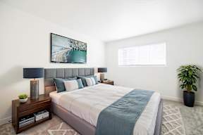 A bedroom with a large bed, a nightstand with a lamp, a potted plant, and a framed picture on the wall.