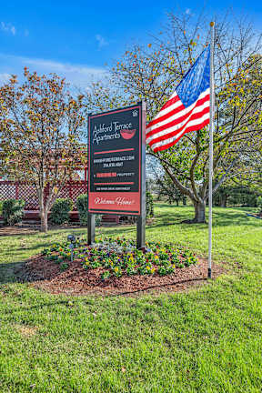 A sign for Ackford Terrace Apartments is in front of a tree and an American flag.