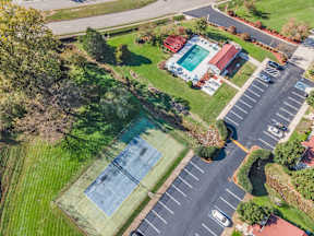 A tennis court is surrounded by a parking lot and a grassy area.