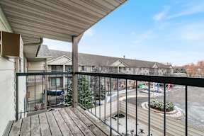 A balcony with a black railing and a wooden floor overlooks a parking lot and other houses.