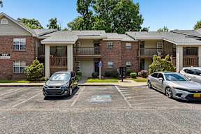 A black car is parked in a parking lot in front of a brick apartment building.