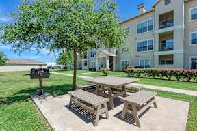 A wooden picnic table is in the foreground of a grassy area with apartment buildings in the background.
