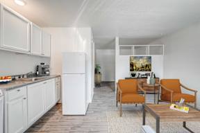 A kitchen with white cabinets and a white refrigerator with a dining area with orange chairs and a painting on the wall.