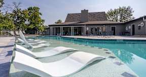 A pool with a white lounger in the foreground and a house in the background.