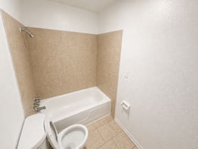 A white toilet is in a bathroom with a white tub located at the pointe apartments in Pasadena, Tx