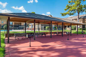 A covered picnic area with tables and benches and the pool in the background at Stone Canyon apartments in Shreveport, LA.