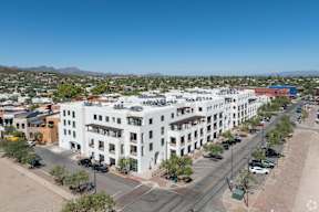 A large white building with balconies is in the middle of a street with cars and trees.