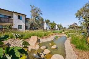 A stream runs through a rocky landscape in front of apartment buildings.