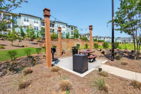 a picnic table and grill sit in a park with a brick wall and apartment buildings in the