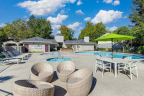 Monte Bello Swimming pool deck with round wicker-like lounge chairs.  There is a square table, chairs and line green umbrella for additional seating around the swimming pool. at Monte Bello Apartments