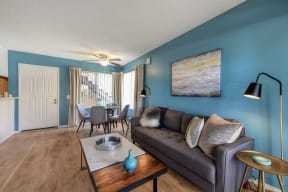 Model home living room entrance and sitting area with hardwood inspired flooring, couch, coffee table, and dining area in the corner.  There is a light blue accent wall. 