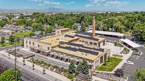A large building with a tall chimney is surrounded by a parking lot and other buildings.