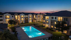 A swimming pool is surrounded by a grassy area and apartment buildings.