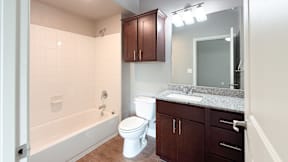 A white toilet sits in a bathroom with a white tub and brown cabinets.