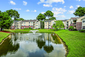 A pond with a fountain in the middle of a grassy area in front of apartment buildings.