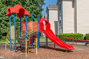Red Yellow and Blue Playground with Red Seesaw with Building Exterior and Treeline in the Background
