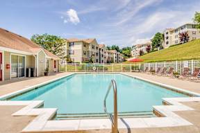 Swimming pool with lounge chairs alongside the pool. Tress and apartment buildings in the background