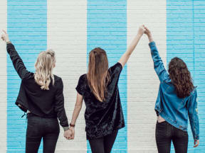 Three female friends standing facing a painted white and baby blue brick wall holding hands in the air in celebration
