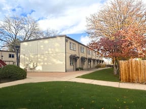 A building with a grey facade is surrounded by a green lawn and a wooden fence.