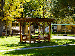 A wooden pergola with benches is situated in a grassy area with trees at Garden Court Apartments, Denver, CO