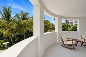 A white room with a wooden chair and table with a view of palm trees outside.