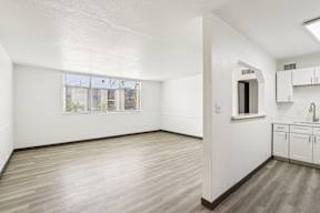 A room with white walls and wooden flooring, with a window and a small kitchenette.