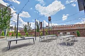 A patio with tables and benches is set up with a brick wall and a water tower in the background.