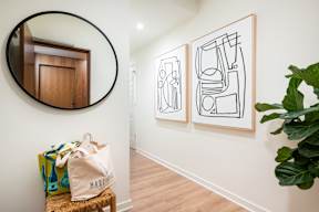 A room with a round mirror, a bag on a stool, and two framed artworks on the wall.