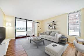 A living room with a grey couch, a grey ottoman, a grey rug, a grey coffee table, a white chair, a white side table, a white TV, a white wall, a window, a door, a balcony