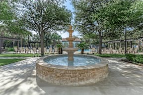 A fountain in the middle of a park with trees and chairs around it.