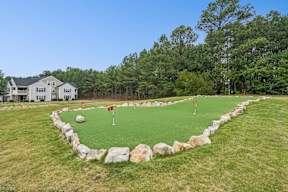 A small putting green is surrounded by rocks and grass in front of a house.