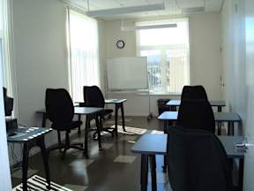 A classroom with black chairs and tables, a whiteboard, and a clock on the wall.
