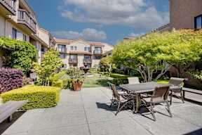 A patio with a table and chairs is surrounded by green plants and trees.