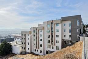 A modern building with a mix of white and dark grey exterior is situated on a hill with a clear sky in the background.