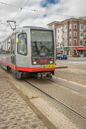 A red and silver train with the words "Do Not Pass" on the back is on tracks in front of a building.