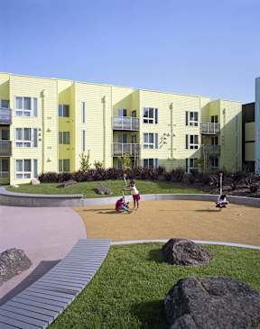 Two children playing in a sandbox in front of a yellow building.