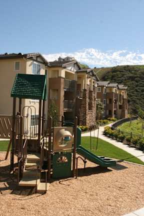 A playground with a green slide and a seesaw in front of apartment buildings.