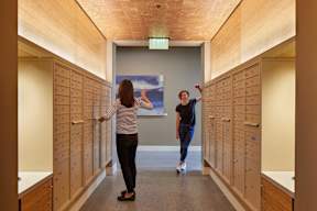 Two people walking down a hallway with wooden lockers on either side.