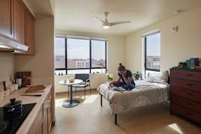 A woman is sitting on a bed in a room with a kitchen area.