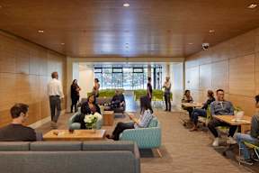 A group of people are sitting and standing in a room with wooden walls and a large window.